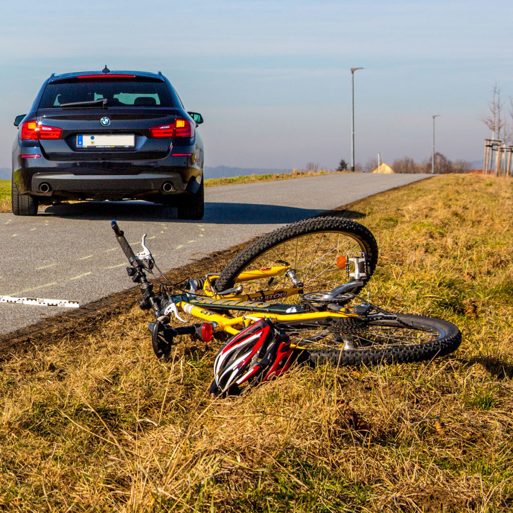 Im Vordergrund liegt ein Fahrrad zusammen mit einem Fahrradhelm neben der Straße auf dem Rasen, ein Stück weiter hinten auf der Straße steht leicht schräg ein Auto mit eingeschaltetem Warnblinklicht